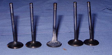 Five exhaust valves laid side by side: one stock straight 944 Turbo valve in the center flanked by four bent valves removed from the damaged head